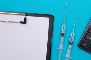 Vaccination, Immunology or Revaccination Concept - Two Medical Syringe Lying on Blue Table in Doctor's Office in a Hospital or Clinic. Black Clipboard with Sheet of Paper - Mock Up with Copy Space photo