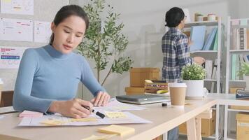 Two young Asian startup partners discuss and brainstorm with memo note in casual small business office, marketing planning jobs on desk, and freelance work. E-commerce entrepreneur and coworker team. video