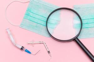 Anti Vaccine Movement and Anti Mask Concept - Broken Medical Syringe and Green Torn Medical Face Mask with Magnifying Glass on the Pink Background. Pandemic and Lockdown End - Top View, Flat Lay photo