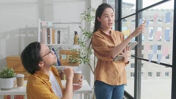 Two young Asian startup company partners discuss and brainstorm, memo notes at casual small business office windows, marketing planning jobs, and freelance work. E-commerce entrepreneur coworker team. video