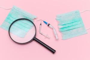 Anti Vaccine Movement and Anti Mask Concept - Broken Medical Syringe and Green Torn Medical Face Mask with Magnifying Glass on the Pink Background. Pandemic and Lockdown End - Top View, Flat Lay photo