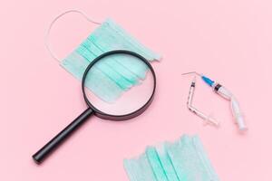 Anti Vaccine Movement and Anti Mask Concept - Broken Medical Syringe and Green Torn Medical Face Mask with Magnifying Glass on the Pink Background. Pandemic and Lockdown End - Top View, Flat Lay photo