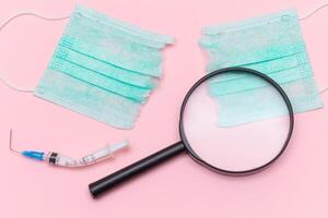Anti Vaccine Movement and Anti Mask Concept - Broken Medical Syringe and Green Torn Medical Face Mask with Magnifying Glass on the Pink Background. Pandemic and Lockdown End - Top View, Flat Lay photo