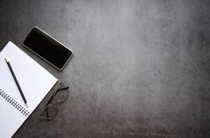Flat lay top view notebook and pencil with mobile phone and glasses on light black color office desk photo