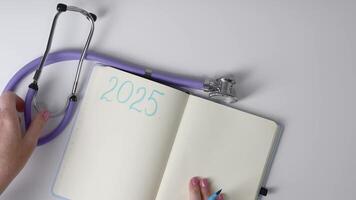 fêmea médico escrevendo dentro caderno metas para 2025 escrito em página deitado em branco fundo Próximo para estetoscópio, conceito do configuração saúde relacionado metas para futuro, video