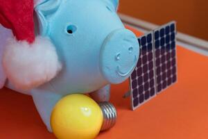 Piggy bank coins with Santa Claus christmas hat, photovoltaic solar panel and lamp isolated on orange background. Concept image. photo