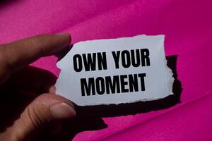 Own your moment message written on ripped torn paper piece with a pink background. Conceptual own your moment symbol. Copy space. photo