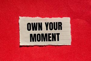 Own your moment message written on ripped torn paper piece with red background. Conceptual own your moment symbol. Copy space. photo