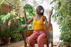 alegre negro mujer bailando sentado en silla escuchando favorito música rodear por exótico plantas de interior foto