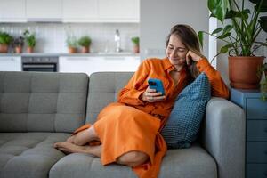 contento mujer utilizando Teléfono móvil a hogar sofá. satisfecho relajado hembra tener Internet divertido, chateando en línea. foto