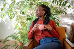 soñando africano americano mujer participación taza descansando en Sillón rodear plantas de interior mirando aparte. foto