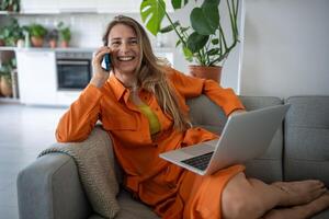 contento relajado mujer sonriente hablando en teléfono inteligente sentado con ordenador portátil en sofá tener agradable diálogo foto