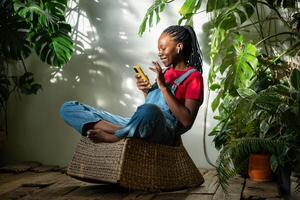 alegre negro mujer relajante a interior jardín. africano americano con teléfono inteligente entre soleado jardín foto
