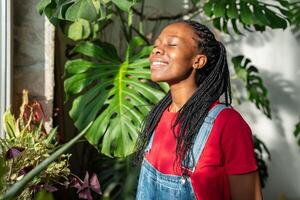 sonriente negro mujer entre interior jardín. africano americano contento alegre niña en contra de planta hojas foto