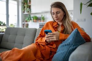 calma enfocado europeo mujer utilizar Teléfono móvil sentado en sofá haciendo en línea compras o orden entrega. foto
