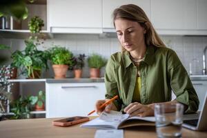 Focused woman attentive budget sitting at table write down expenses in notebook use phone calculator photo