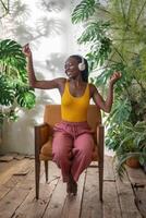 alegre negro mujer bailando sentado en silla escuchando favorito música rodear por exótico plantas de interior foto