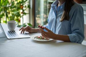 Peaceful woman at table in morning holding sandwich having breakfast using laptop computer. photo