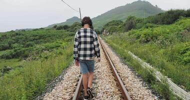 Teenager in plaid shirt and denim shorts walking alone on railway tracks in a green countryside, captured from the back with themes of solitude and introspective journey video
