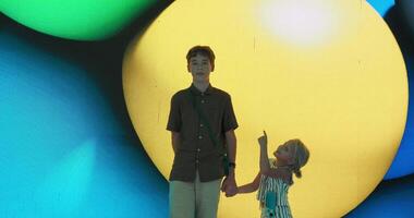 Brother and sister stand holding hands while posing for photos as the colorful background behind them changes video