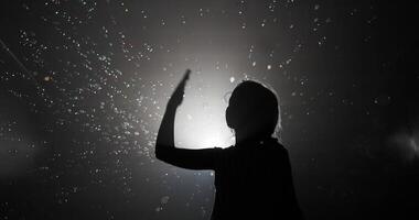 Silhouetted view of a child extending an arm to pop soap bubbles in a mist-filled room, illuminated by a bright light, creating a playful and mysterious ambiance video