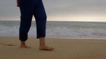 Close-up of a person walking barefoot along a sandy beach at sunset, with ocean waves in the background. The person's face is not visible, only legs and feet captured video