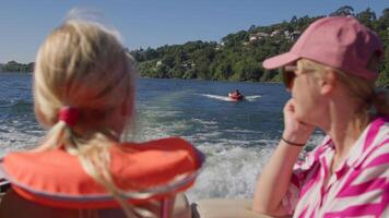 Two children with life jackets on boat watching a person tubing on a lake, surrounded by trees and hills, shot from behind video