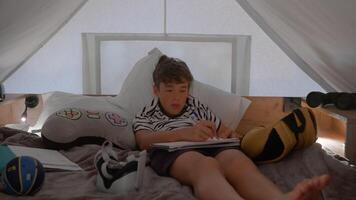 Teen boy in striped shirt writing in a notebook while relaxing inside a glamping tent. The tent is cozy, with various personal items around him. He appears focused on his task video