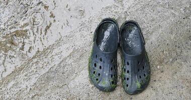 A pair of black rubber clogs with green grass stuck on them, resting on wet concrete pavement during a rainy day. The surface is covered in water, enhancing the texture of the concrete video
