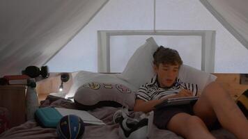 A teenage boy is lying in a glamping tent, writing in a notebook with various personal items around him. The cozy setting shows elements of comfort and relaxation video