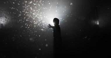Silhouetted view of a single child leaning forward, reaching out to pop soap bubbles in a misty room against a bright light source, creating a playful and mysterious scene video