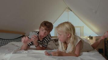 Caucasian brother and sister lying on bed in a tent, engaged in playing a card game, expressing concentration and enjoying each other company in a calm setting video