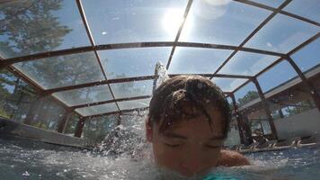 Close-up of a young boy underwater in a swimming pool, with sunlight streaming through a glass roof above and water droplets creating a sparkling effect around him video