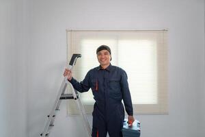 Smiling Worker with Ladder and Toolbox in Front of Window Blinds photo