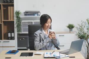 Successful Asian businesswoman smiling using laptop computer and holding coffee cup at office. Confident Asia businesswoman sitting happily in the office. photo