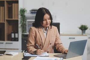 Asian Businesswoman Using laptop computer and working at office with calculator document on desk, doing planning analyzing the financial report, business plan investment, finance analysis concept. photo