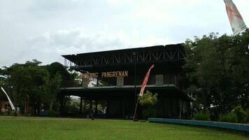Banyumas, Indonesia - November 5 2024, A modern building labeled ANDHANG PANGRENAN surrounded by lush greenery and open grass, under a cloudy sky with colorful flags. video
