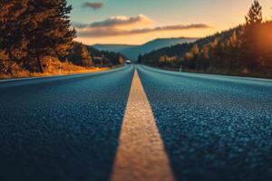 A long, straight asphalt road stretches into a hazy sunset with a yellow dividing line and forested mountains in the distance. photo