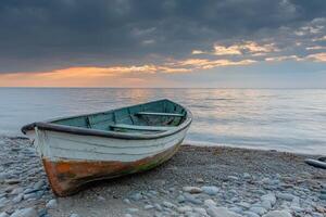 A weathered boat sits on a pebbled shore with a dramatic sunset in the background. photo