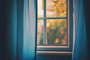 A close up of a window with blue curtains partially drawn, showing a view of a blurry green tree. photo