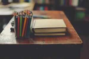 A wooden desk with a stack of books, a container of colored pencils, and a blank piece of paper. photo