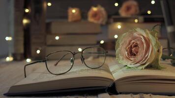 A tranquil reading nook features an open book resting on a table, accompanied by stylish glasses and a delicate pink rose, all softly illuminated by fairy lights creating a warm atmosphere. video