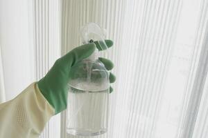 A hand gripping a cleaning spray bottle while wearing a protective glove, positioned by a window photo