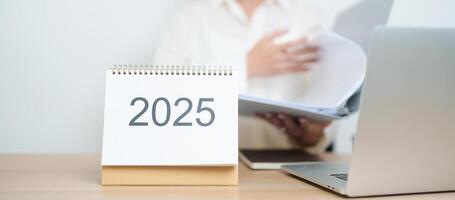2025 Year Calendar on table with business woman using laptop computer, working and writing in office. countdown, Happy New Year, Resolution, Year End and New Start and Goals Plan Action Concept photo