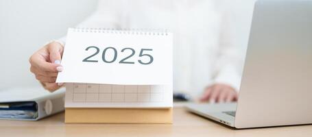 2025 Year Calendar on table with business woman using laptop computer, working and writing in office. countdown, Happy New Year, Resolution, Year End and New Start and Goals Plan Action Concept photo