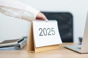 Business woman change 2025 Year Calendar on table with laptop computer during working and writing in office. countdown, Happy New Year, Resolution, Year End and New Start and Goals Plan Action Concept photo