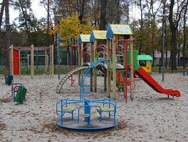 Sand covered with autumn leaves on the empty multi-colored playground with slides and small merry go round. No children at cold season at the play yard photo