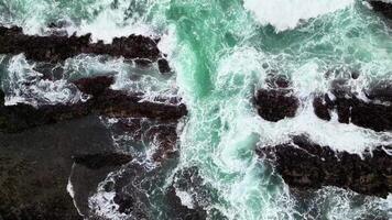 Aerial top view the ocean waves crash against the black rocks, creating white foam that contrasts with the dark green seawater. The water flow appears strong, breaking through the rough rock crevices. video