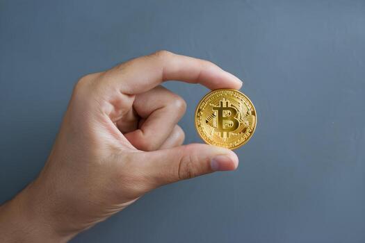 Hand holds golden Bitcoin coin between thumb and index finger, blue background photo