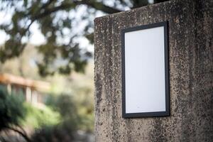 White signboard with black frame mounted on concrete wall, trees slightly blurred behind photo